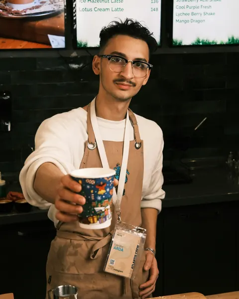 Barista mit Schürze reicht einen bunt bedruckten Kaffeebecher über den Tresen